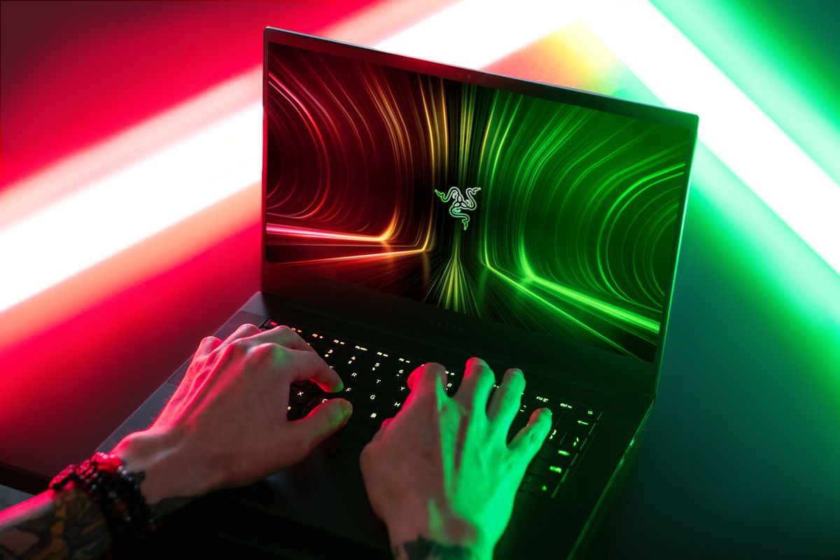 Toma por encima del hombro de un usuario que escribe en un Razer Blade 14. La pantalla muestra el logotipo de Razer sobre un fondo rojo, verde y negro.